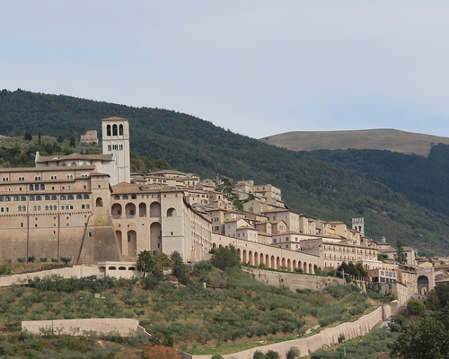 Weitblick auf die mittelalterliche Stadt Assisi mit der imposanten Basilika San Francesco am Hang des Monte Subasio, umgeben von grüner Hügellandschaft.